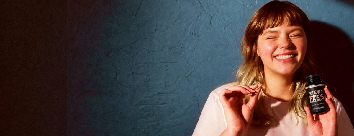 A laughing woman holding a Potency Fresh capsule and bottle against a blue wall